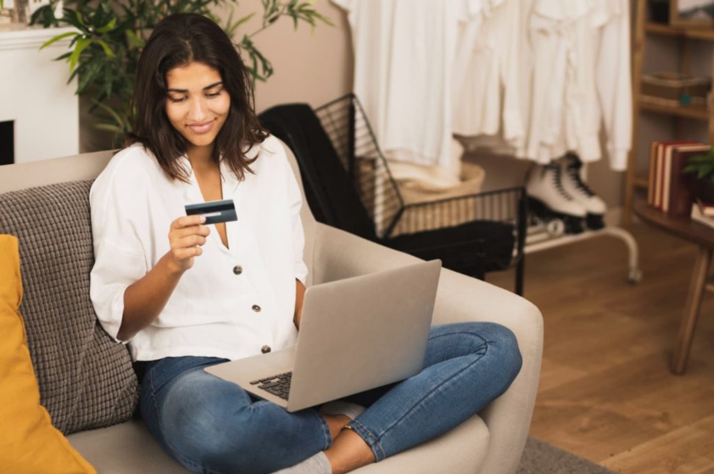 A young woman sitting on a couch shopping online with a credit card in hand and a laptop on her lap in a home setting.