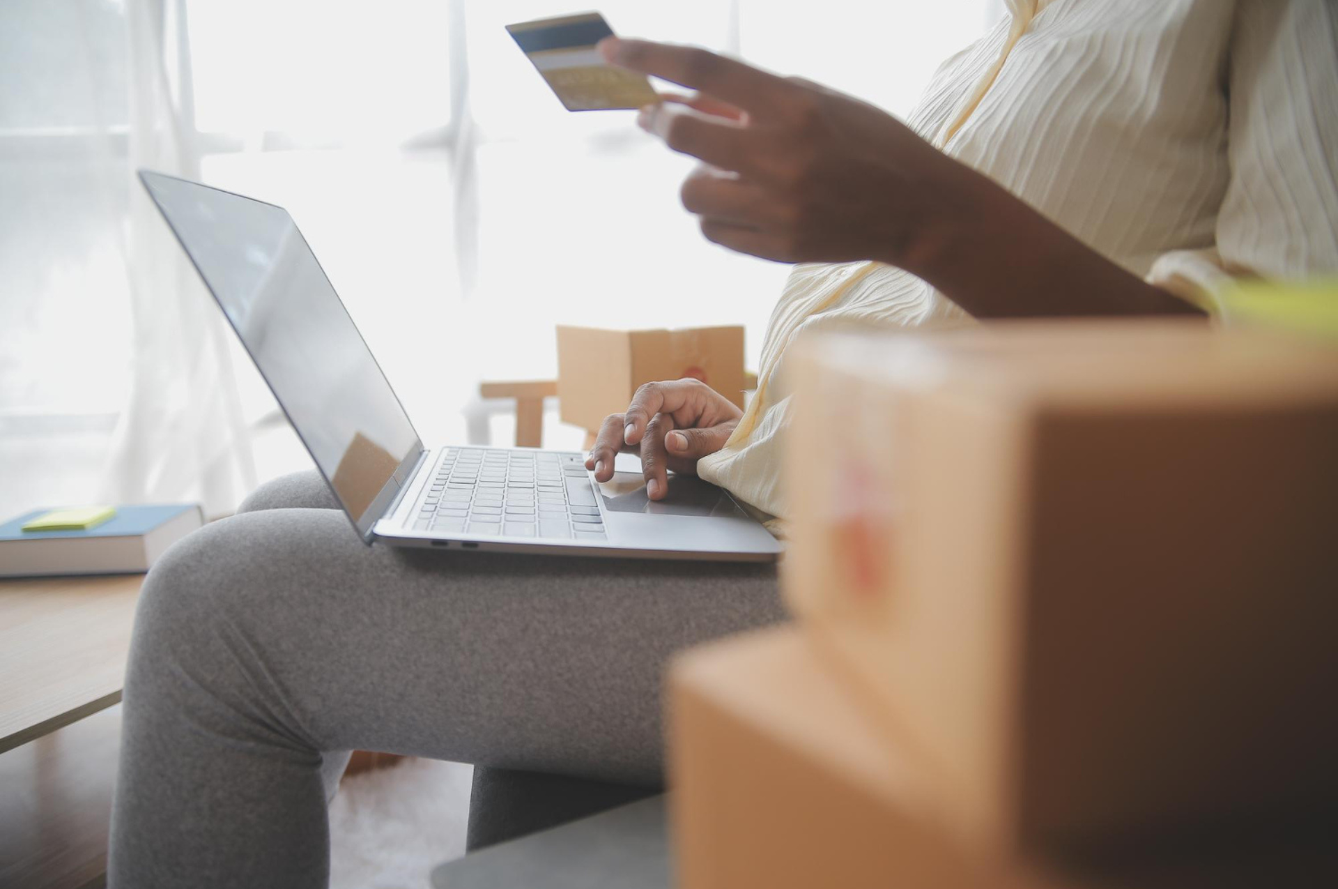 A person seated with a laptop and credit card, surrounded by boxes, managing online sales.