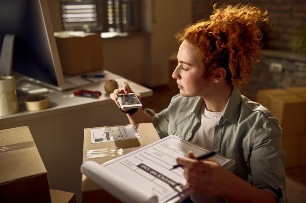 A woman with curly red hair checking an item with a handheld scanner while reviewing a clipboard in a shipping facility, indicating inventory management.