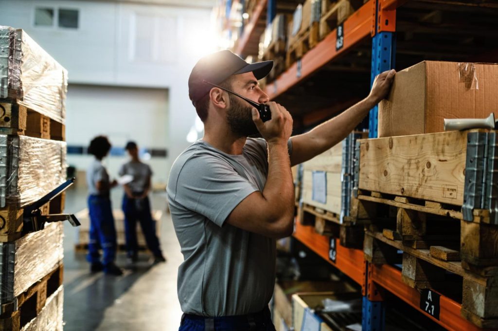 Image depicting a warehouse worker in a cap, communicating via walkie-talkie while placing a box on a shelf